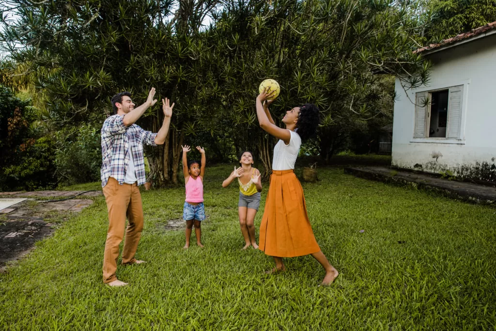 
Família composta por duas crianças e dois adultos, jogando bola no gramado em frente a uma casa no campo. Eles aproveitam a vida ao morar no interior.